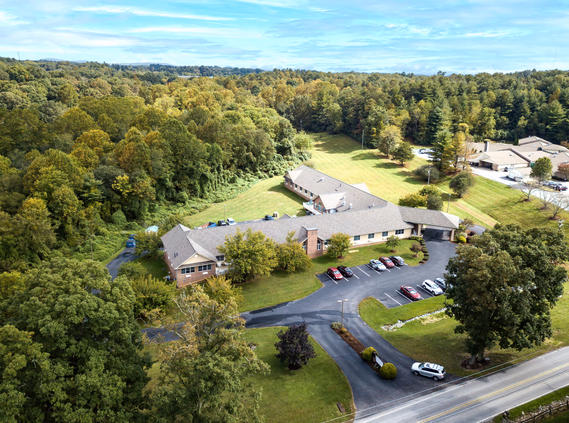 Aerial view of Cherry Springs Village, a large single-story building surrounded by lush green trees and grass. The building has a parking lot with several cars parked, and a road runs along the front of the property. The area is peaceful and scenic with dense forest in the background under a partly cloudy sky.