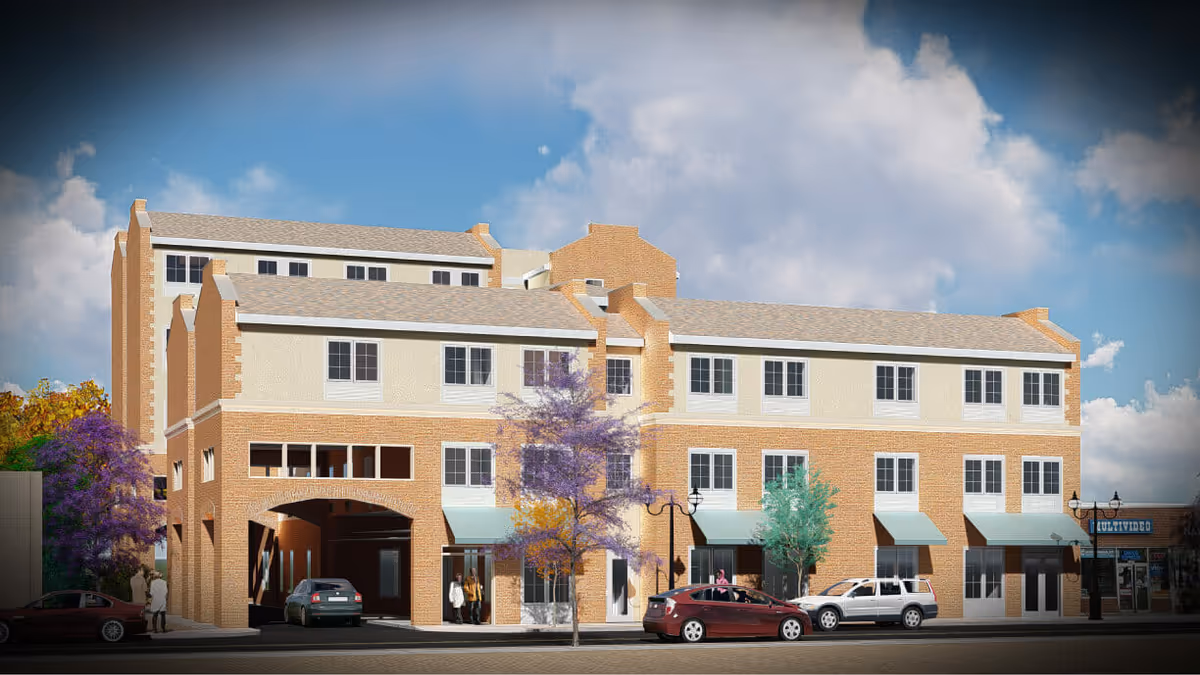 Three-story brick and stucco senior living building with street-level awnings, parked cars and trees under a blue sky.