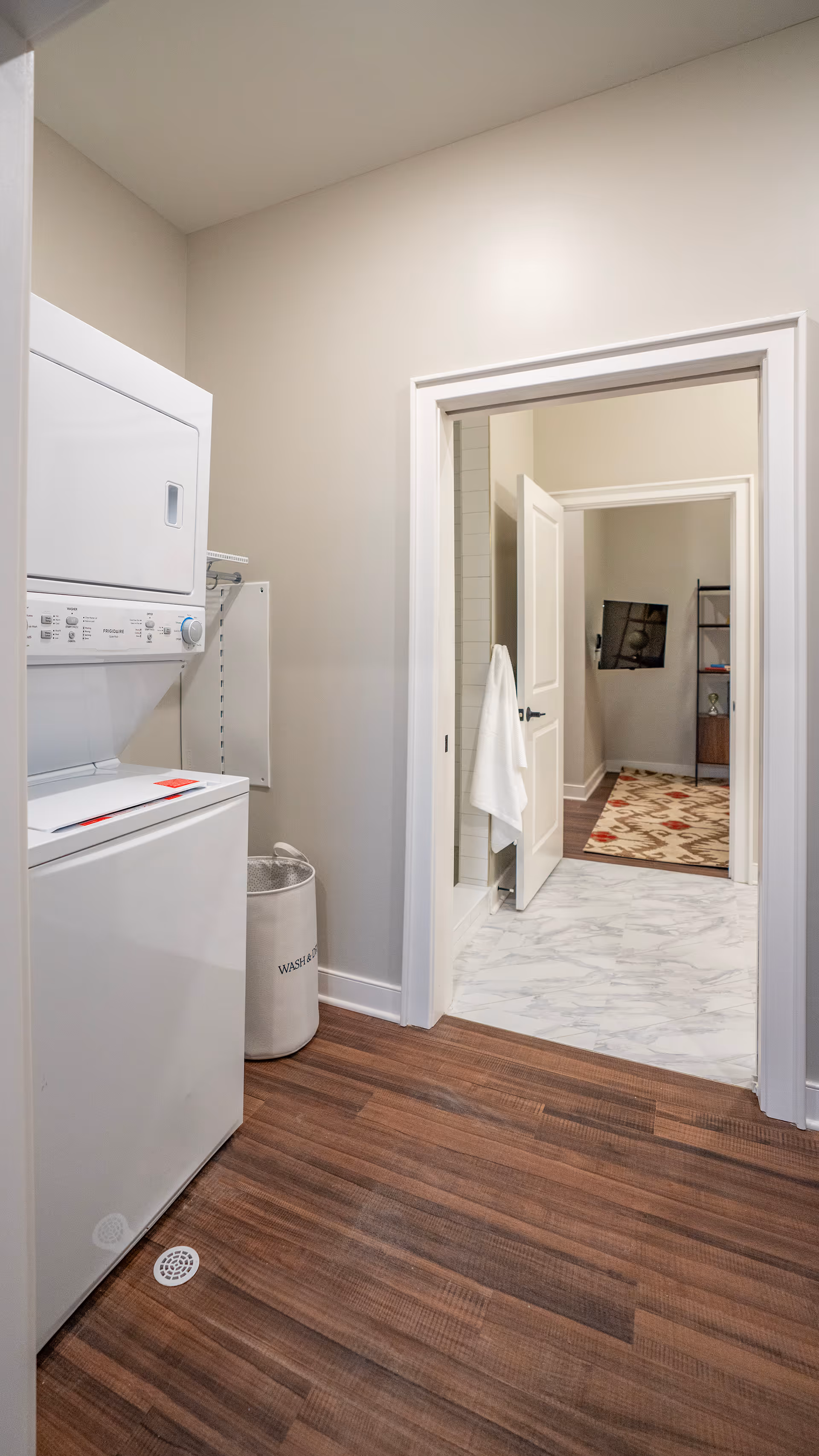 Laundry area with a stacked washer and dryer unit on the left, a laundry hamper labeled 'WASH' next to it, and an open doorway leading to a bathroom with a towel hanging on the door and a glimpse of a room with a patterned rug and shelving unit beyond.