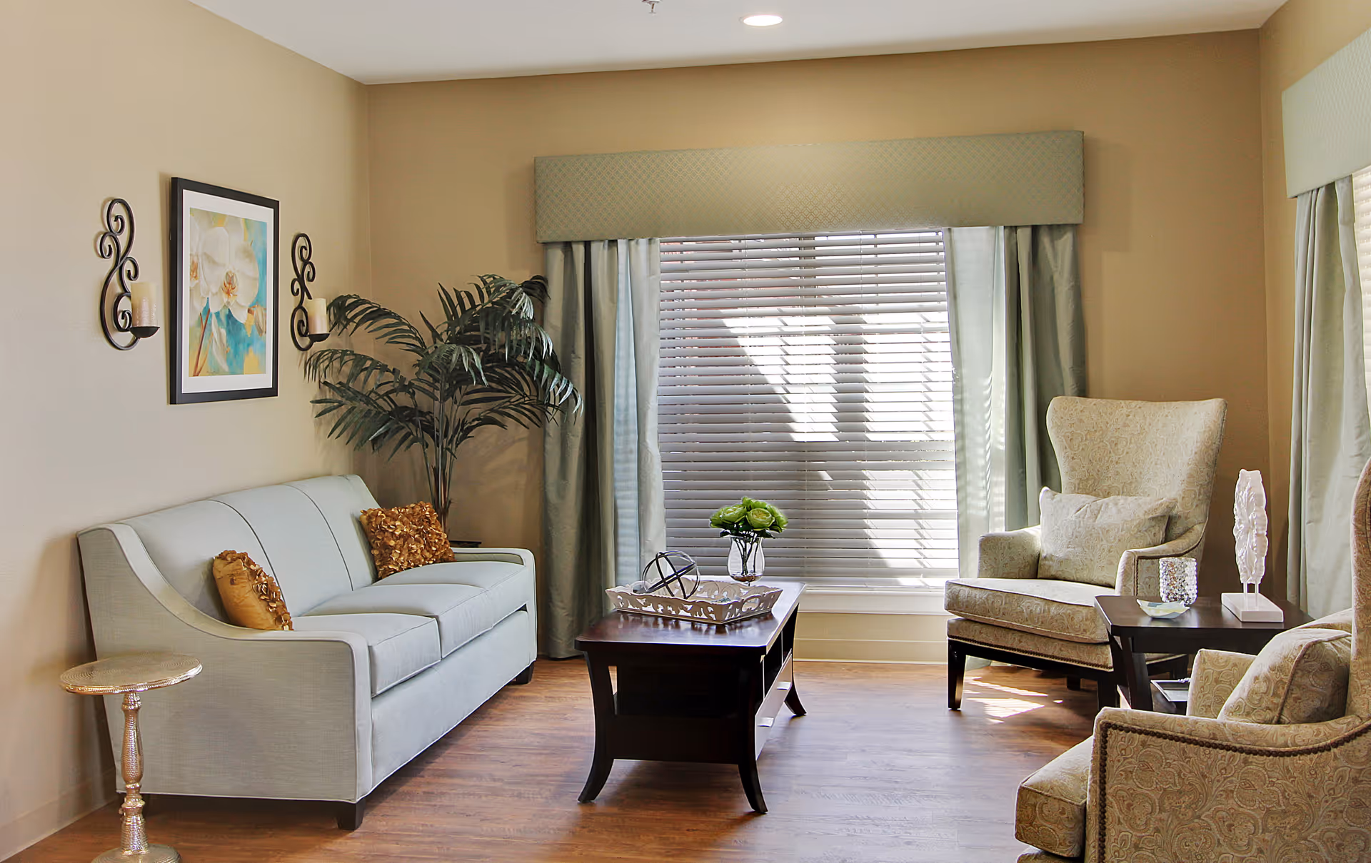 A cozy living room with a light blue sofa adorned with two decorative pillows, two patterned armchairs, a wooden coffee table with a decorative tray and a small vase of flowers, a side table with a decorative item, a large window with blinds and curtains, a potted plant in the corner, and wall decorations including a framed painting and two candle sconces.