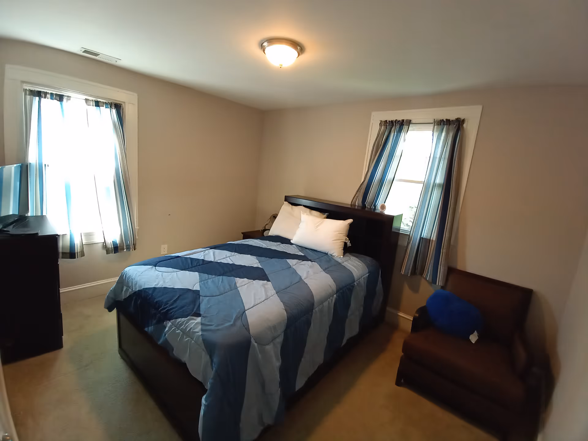A bedroom in an assisted living facility with a bed, dresser, windows, and chair.