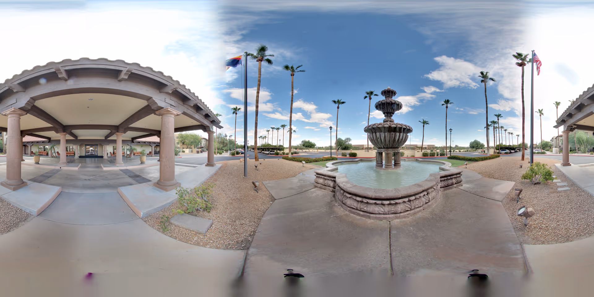 Outdoor view of The Forum at Desert Harbor entrance featuring a covered walkway with columns, a multi-tiered stone fountain with water, palm trees, flags, and a clear blue sky with some clouds.