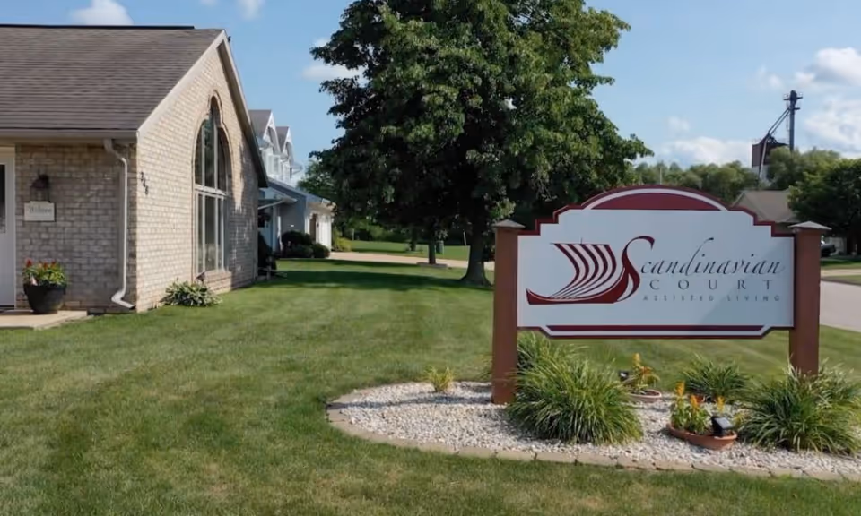 Exterior view of Scandinavian Court Assisted Living facility showing a brick building with a large arched window, a green lawn, trees, and a sign with the facility's name in front.