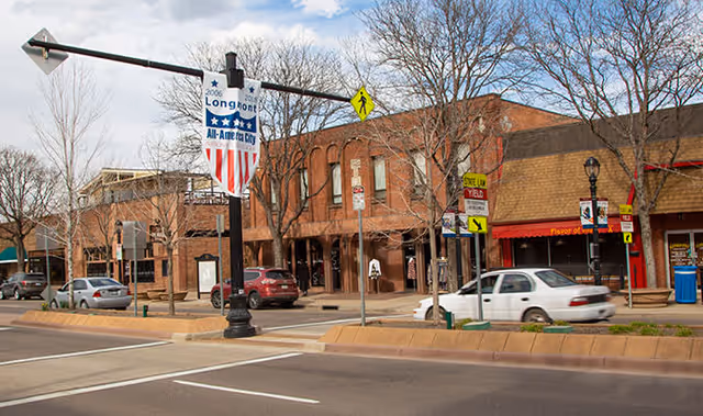 Street view of a small town commercial area with brick buildings, parked cars, leafless trees, and a traffic light pole displaying a banner that reads '2006 Longmont All-America City'.