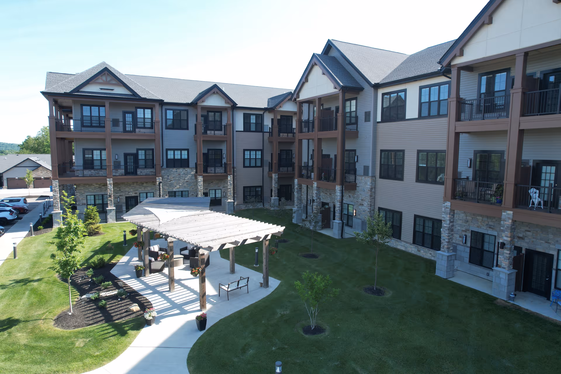Outdoor courtyard area of a multi-story senior living facility with a pergola-covered seating area, green lawn, small trees, and a paved walkway. The building has stone and siding exterior with balconies and large windows.