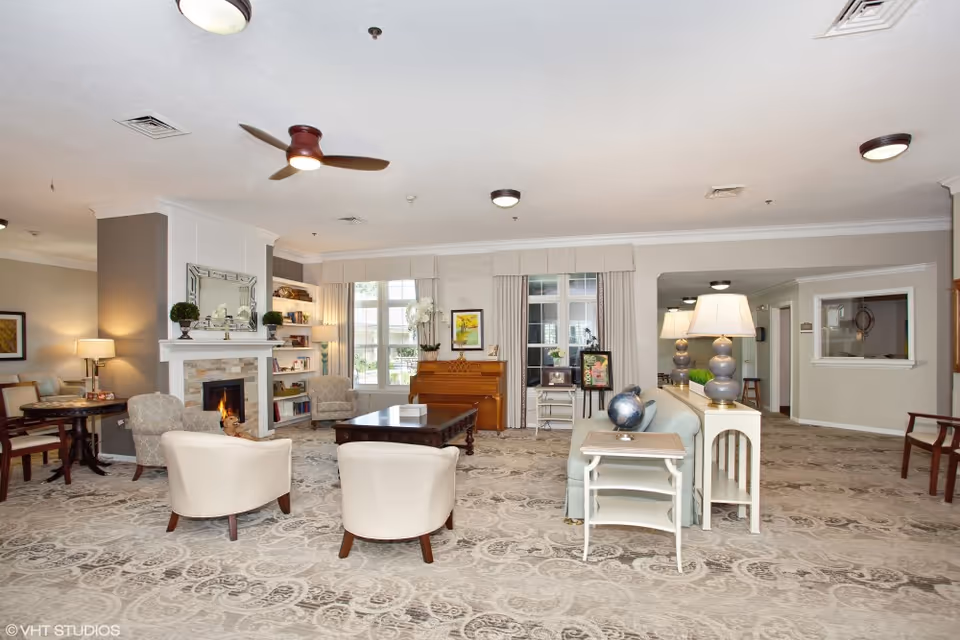 Spacious senior living common room with seating around a fireplace, a piano, and lamps.