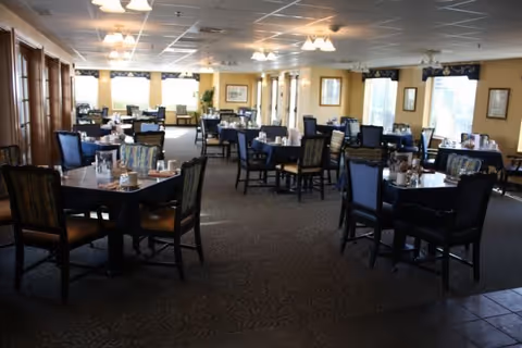 A spacious dining room with multiple tables and chairs arranged neatly. The room has large windows allowing natural light to brighten the space. Each table is set with napkins, glasses, and condiments. The ceiling has several light fixtures, and the walls are decorated with framed pictures and plants.