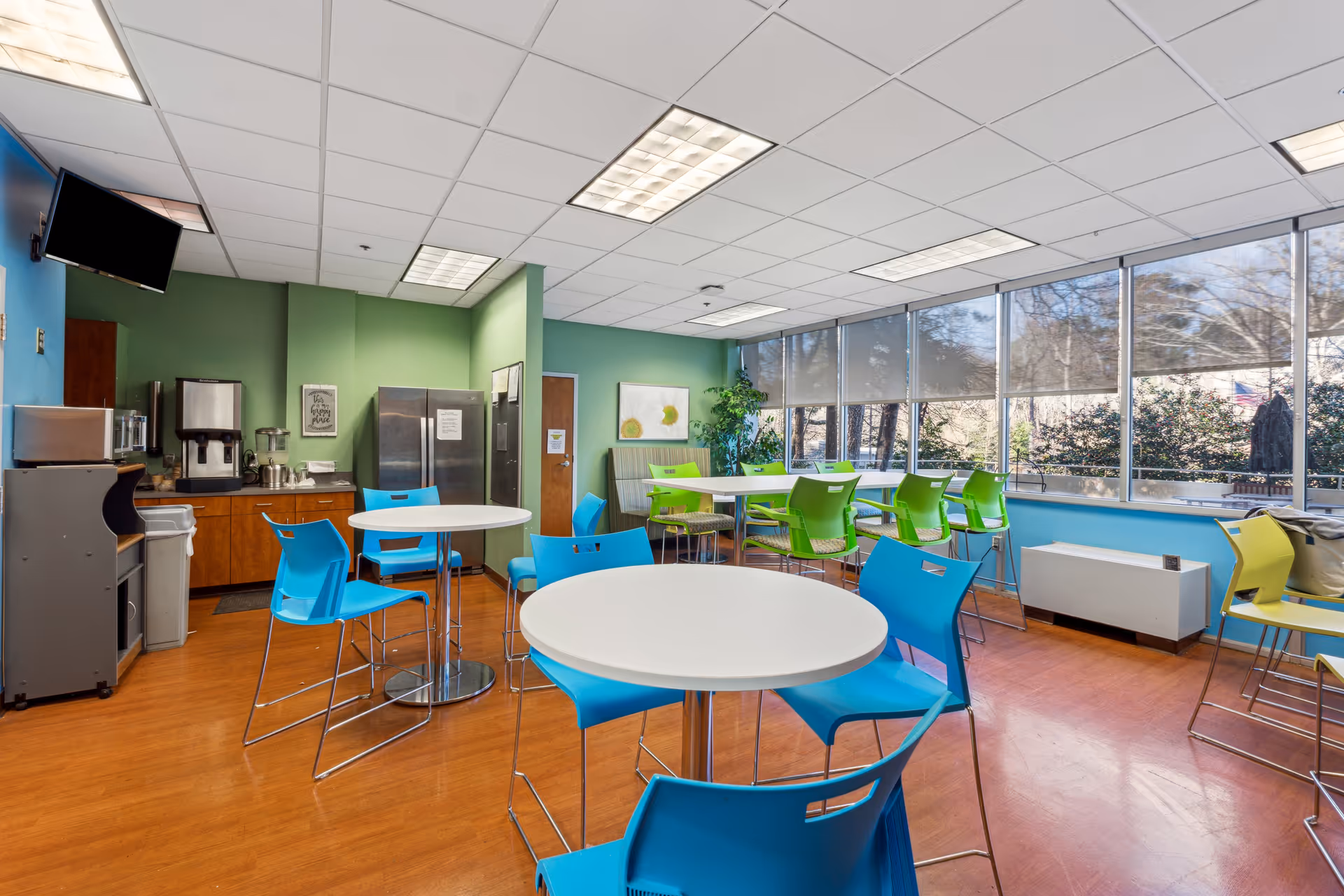 Bright communal dining/break room with round white tables, blue and green chairs, a kitchenette and large windows.
