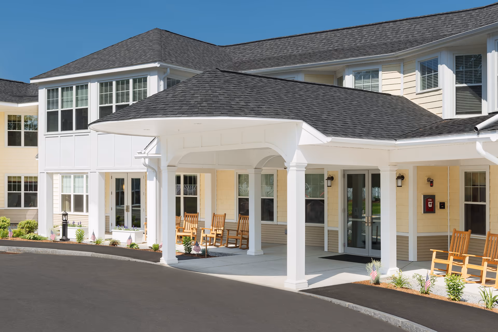 Entrance of a senior living facility with a covered drop-off area supported by white columns, wooden rocking chairs on the porch, beige siding, multiple windows, and a clear blue sky.