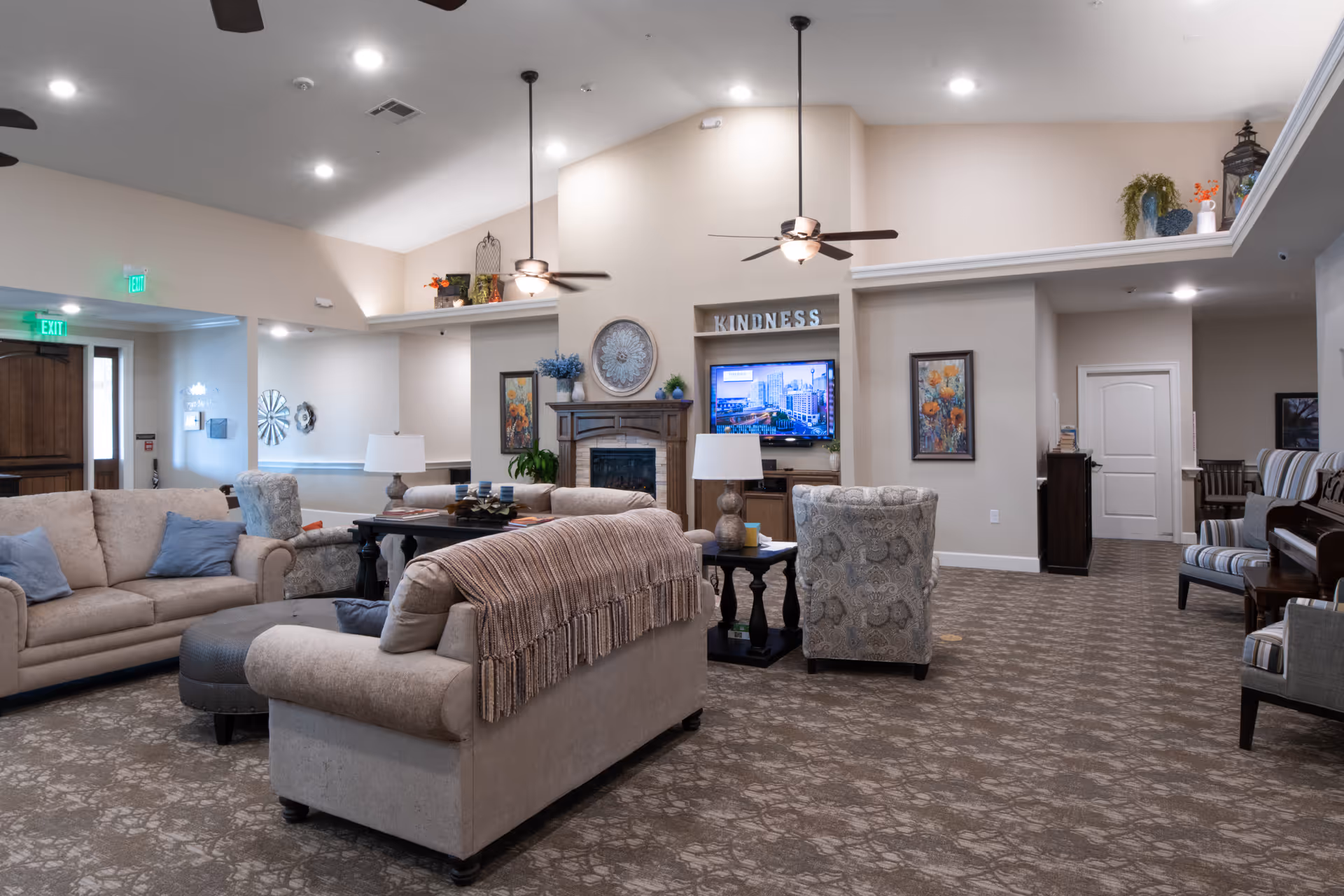 A spacious and well-lit living room area in an assisted living facility featuring beige sofas with blue and beige cushions, a patterned armchair, a fireplace with decorative items, a wall-mounted TV, framed artwork, ceiling fans, and a piano on the right side. The room has carpeted flooring and neutral-colored walls with some plants and decorative pieces on shelves.