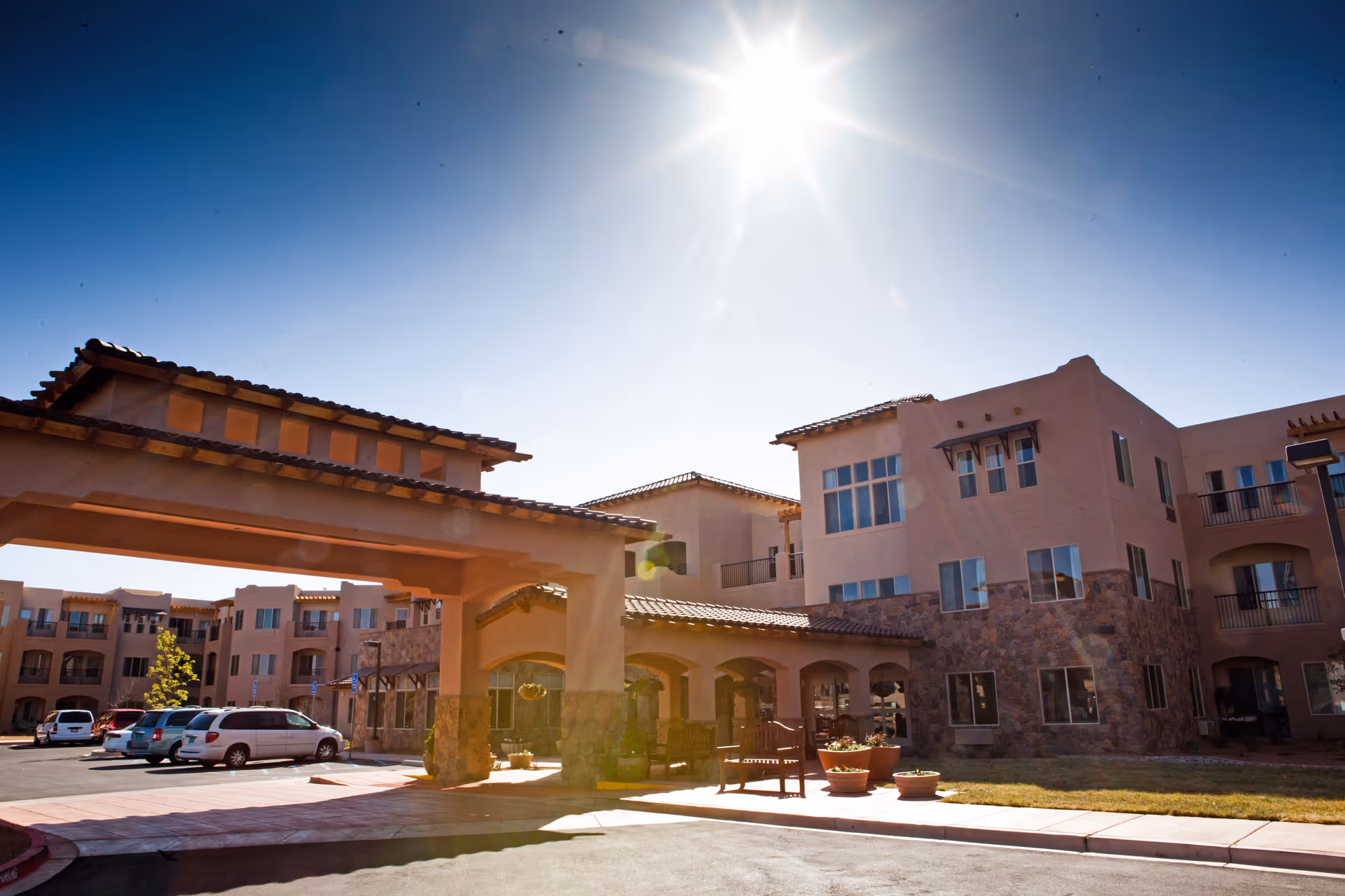 Exterior view of a multi-story retirement living facility under a bright sunny sky with a covered entrance, parked cars, and landscaped surroundings.