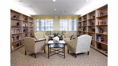 A cozy library or reading room with beige carpet, two large windows with yellow curtains, wooden bookshelves filled with books lining both side walls, and four upholstered armchairs arranged around a small wooden coffee table with a flower arrangement on top.