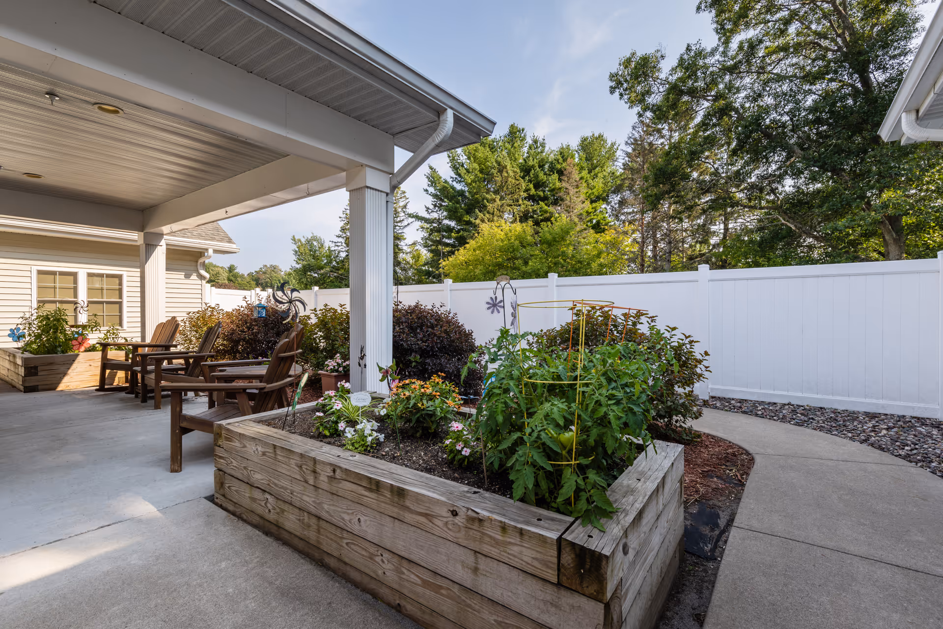 Outdoor patio area at Milestone Senior Living Woodruff featuring wooden raised garden beds with various plants and flowers, several wooden chairs arranged for seating, a concrete pathway, and a white privacy fence with trees in the background under a partly cloudy sky.