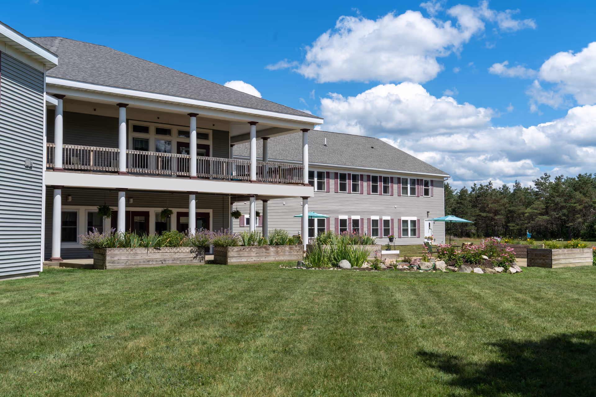 Two-story senior living building with covered porches, white columns, planter beds and a manicured lawn under a blue sky.