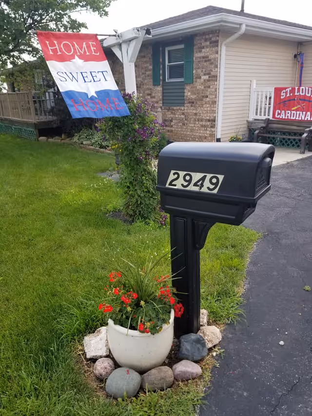 A black mailbox numbered 2949 stands by a driveway in front of a brick-and-siding house with a 'Home Sweet Home' flag and a potted flower arrangement.