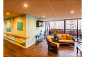 A bright senior living lounge with wicker seating, cushions, colorful wall panels, hardwood floors, and large windows overlooking the city.