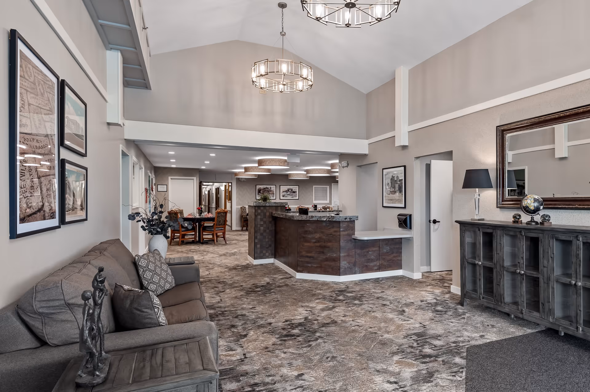 A spacious and well-lit interior common area with a high vaulted ceiling, featuring a gray sofa with decorative pillows and a sculpture on a side table to the left. The room has a carpeted floor, a reception desk in the center with a granite countertop, and a dining area with wooden chairs and tables in the background. The walls are adorned with framed artwork, and there is a large mirror above a cabinet with a lamp and decorative globe on the right side.