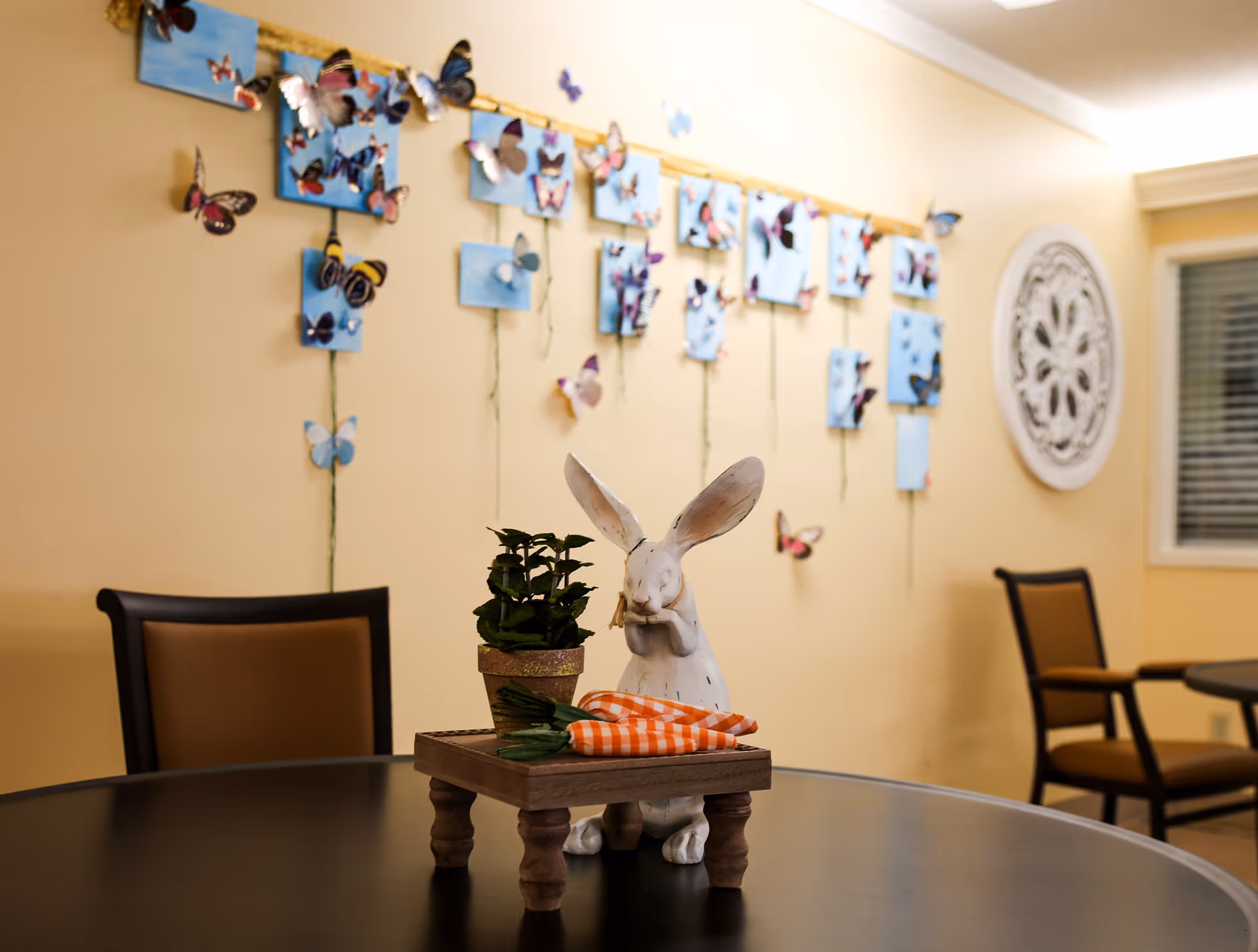A round table with a decorative white rabbit figurine holding an orange and white checkered carrot, a small potted plant, and another carrot on a small wooden platform. In the background, there are two chairs with brown cushions and a wall decorated with blue panels and colorful butterfly decorations, along with a circular white ornamental wall piece and a window with blinds.