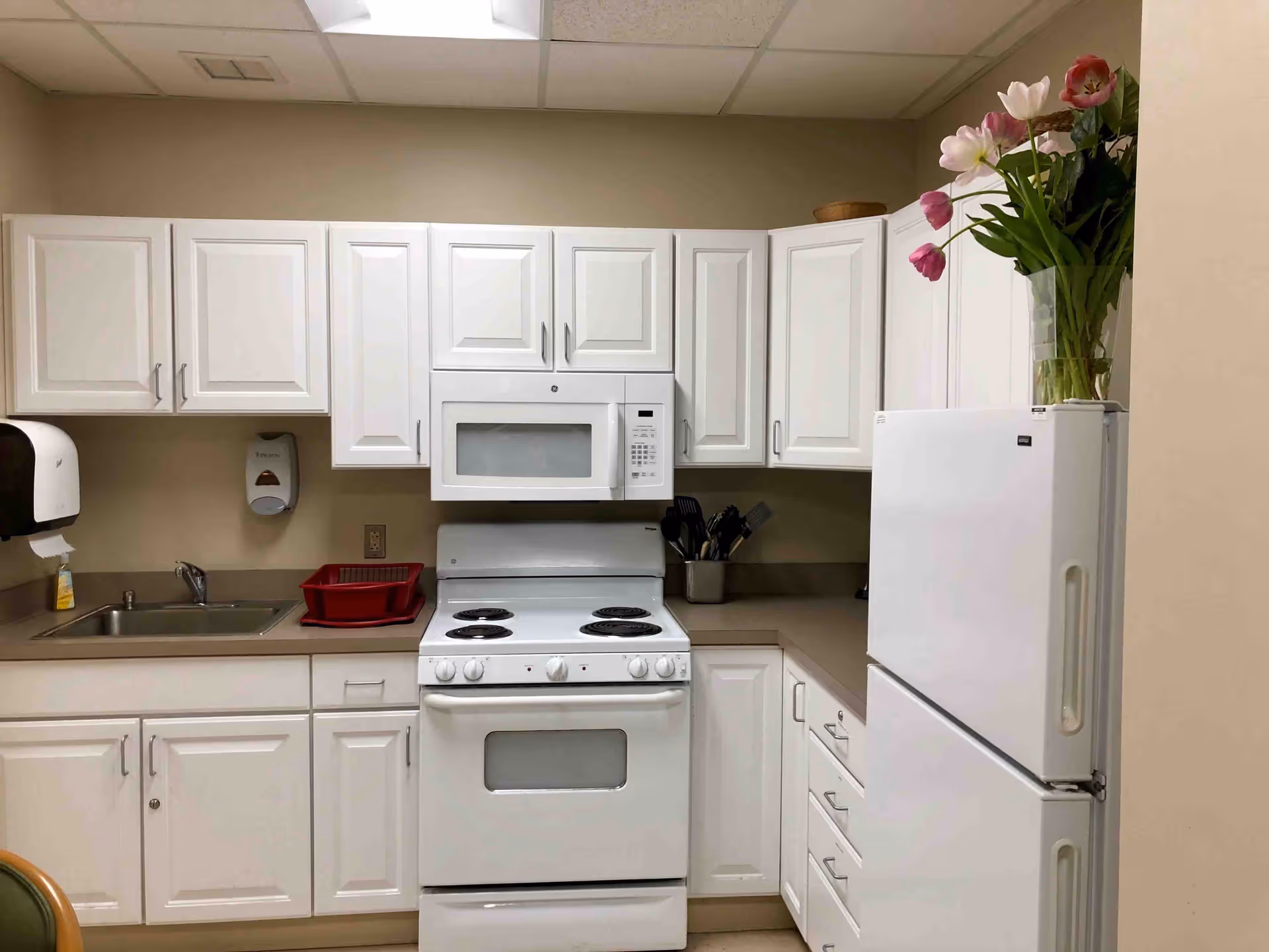 A clean kitchen area with white cabinets, a white electric stove with oven, a white microwave above the stove, a white refrigerator with a vase of pink and white flowers on top, a stainless steel sink with a soap dispenser and paper towel holder on the wall, and a red dish drying rack on the countertop.