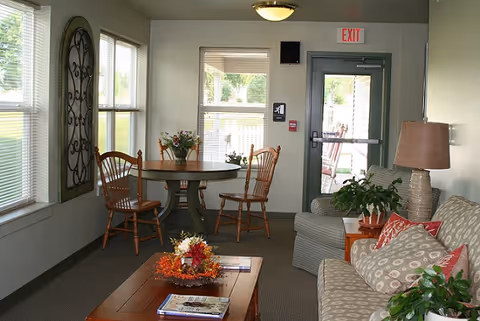 Cozy senior living common room with a sofa, coffee table, round dining table and chairs, plants, and an exit door.