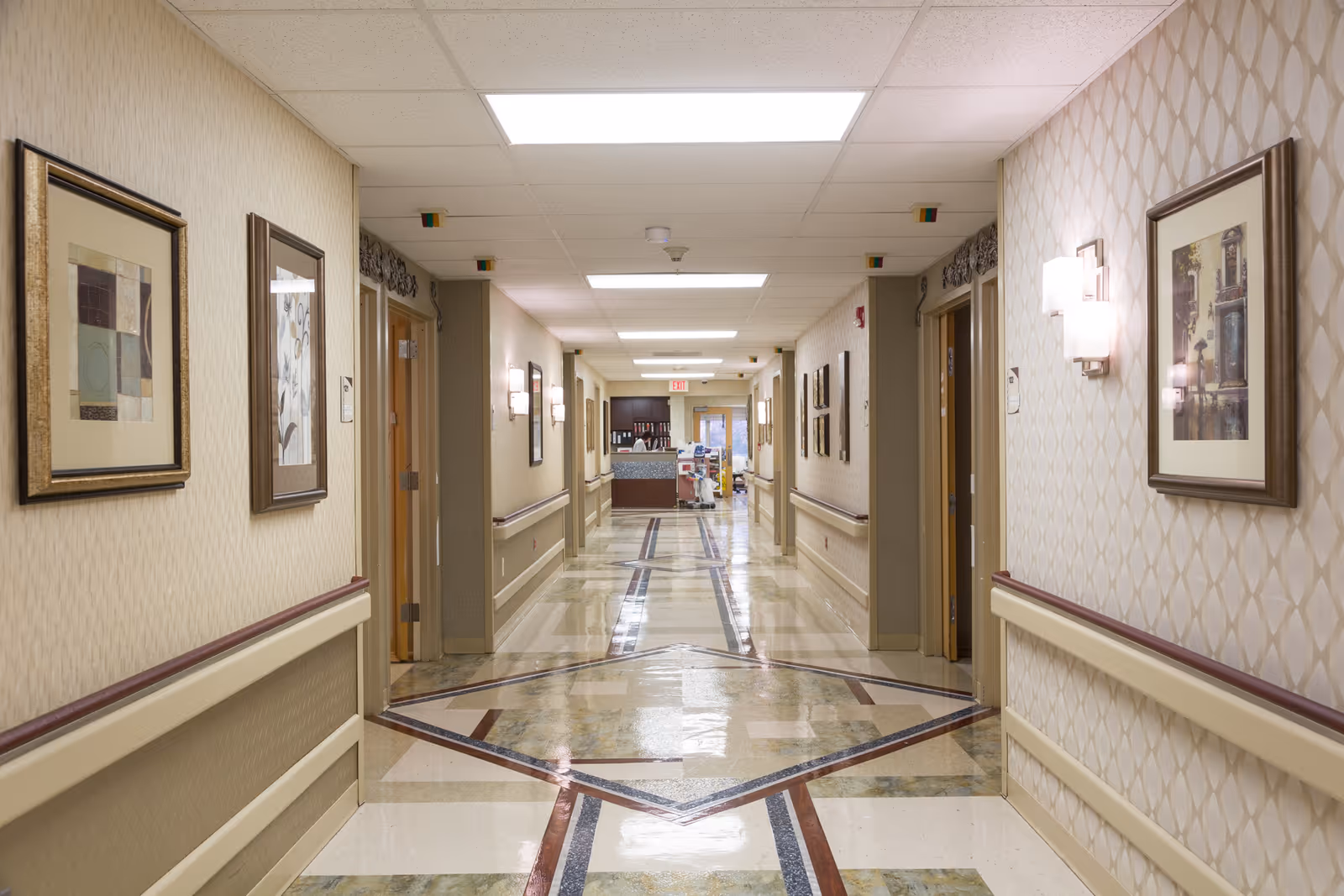 A well-lit hallway in a care center with patterned tile flooring, beige walls adorned with framed artwork, and wall-mounted light fixtures. Doors line both sides of the corridor, and a reception or nurse station is visible at the far end.