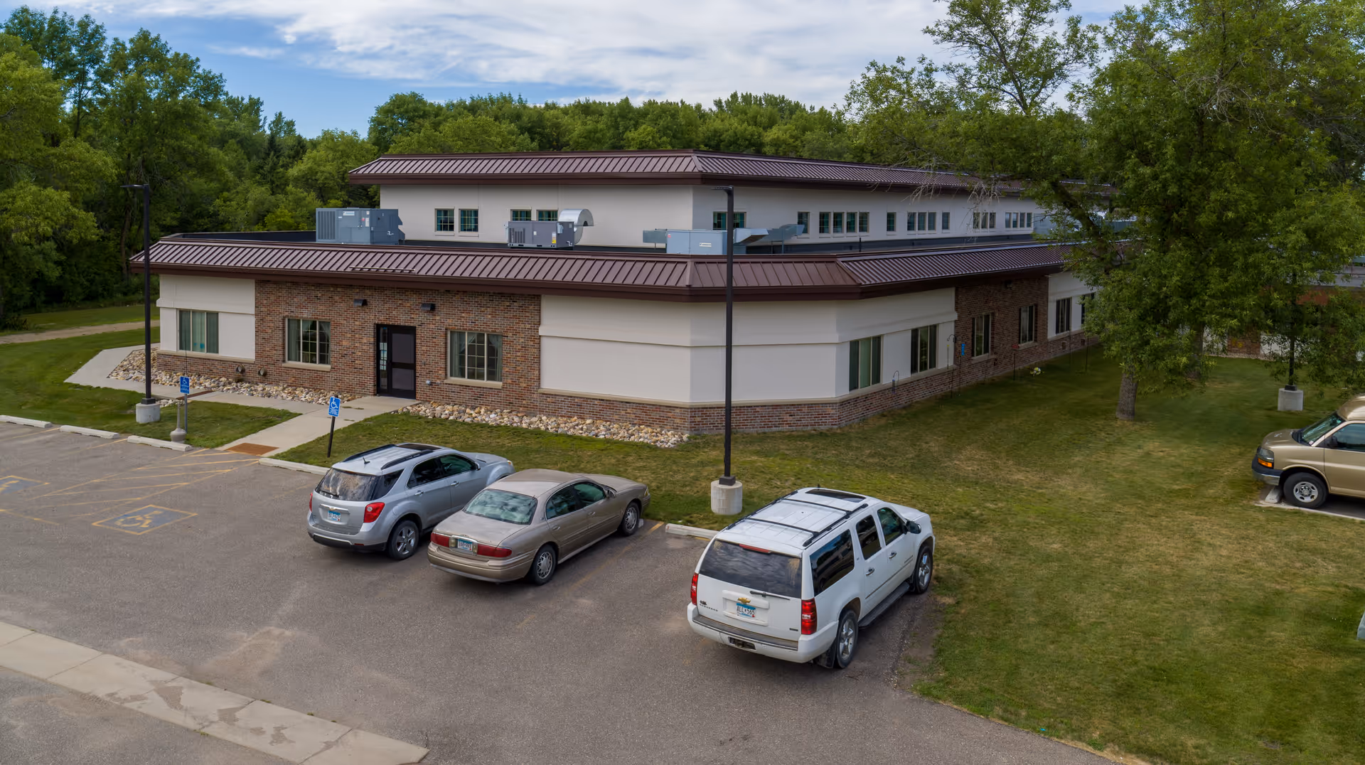 Exterior view of a single-story building with a brick and beige facade, surrounded by green trees and grass. There are four cars parked in front of the building, including a white SUV, a silver hatchback, a beige sedan, and a gold SUV. The sky is partly cloudy.