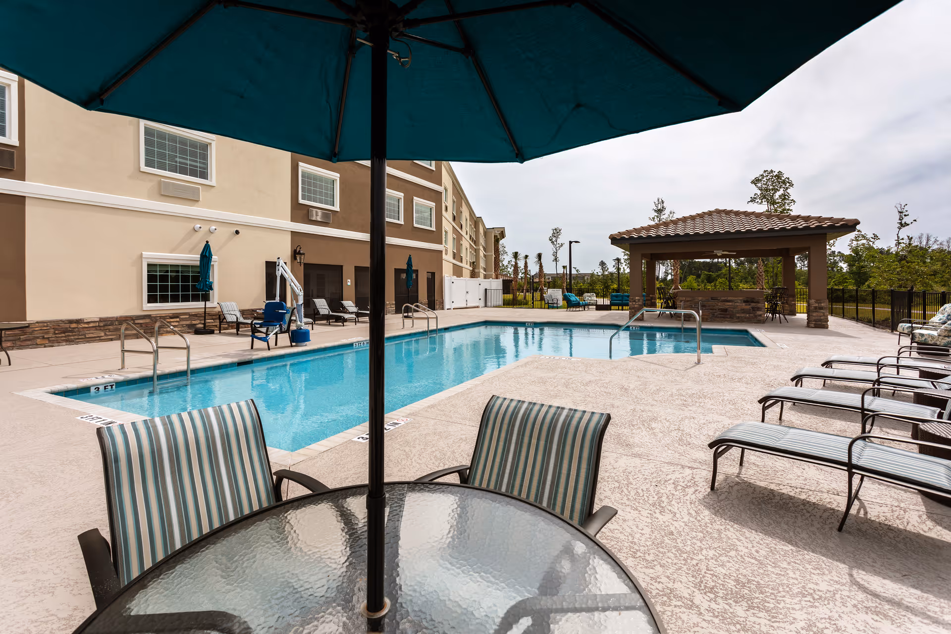 Outdoor swimming pool area at Discovery Village At Deerwood Assisted Living & Memory Care with lounge chairs, a glass table with striped chairs under a large umbrella, and a covered seating area in the background. The pool is adjacent to a multi-story building with windows and doors.