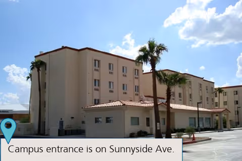 Exterior view of a multi-story beige building with a red-tiled roof, palm trees in front, and a clear blue sky with some clouds above. The building appears to be part of a senior living facility.
