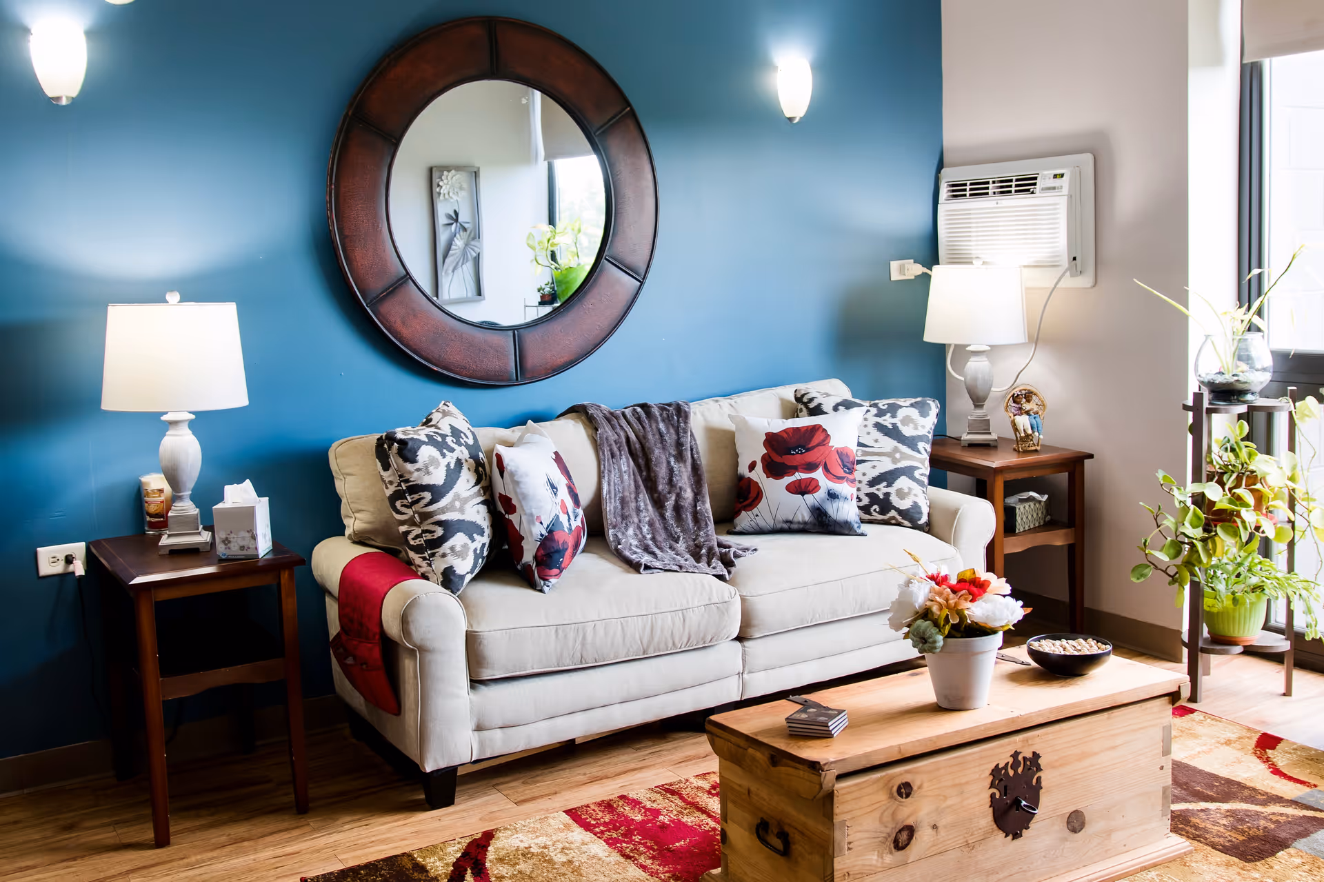 A cozy living room with a beige sofa adorned with decorative pillows and a throw blanket. The wall behind the sofa is painted blue and features a large round mirror. There are two wooden side tables with lamps on either side of the sofa. A wooden chest serves as a coffee table with a flower pot and a bowl on top. A plant stand with green plants is near a window with natural light coming in.