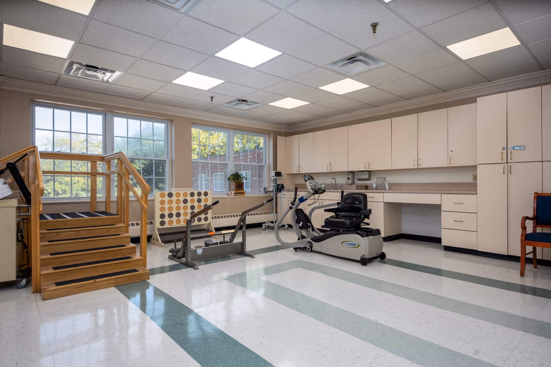Rehabilitation/exercise room with a recumbent exercise bike, parallel bars, wooden practice stairs, cabinets, and large windows.