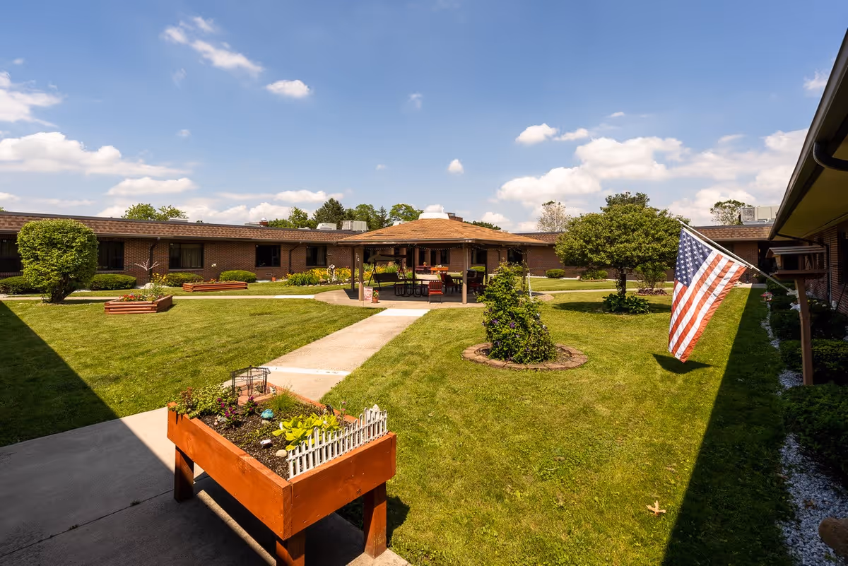 A sunny outdoor courtyard at Wood Haven Healthcare featuring a well-maintained lawn, a raised garden bed with plants, a paved walkway, a gazebo with seating, various shrubs and trees, and an American flag mounted on the building.