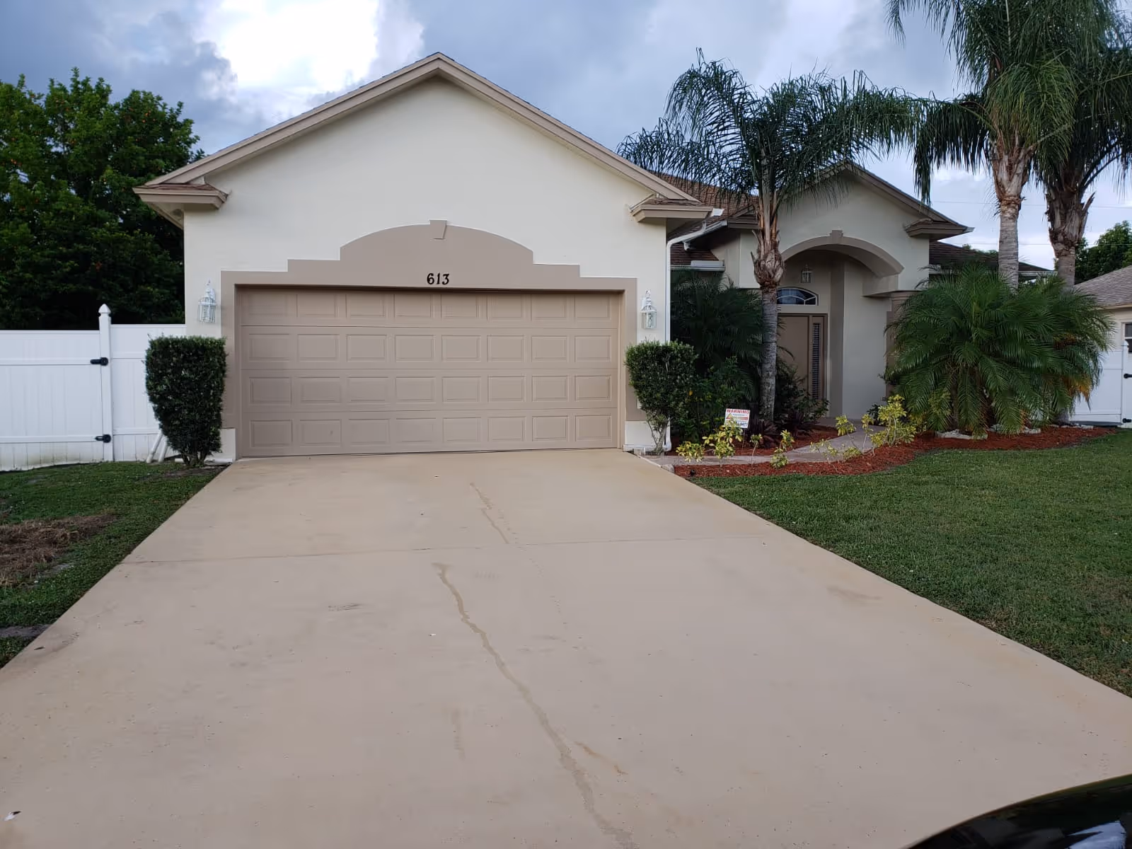 Front exterior view of a single-story house with a beige garage door, a driveway, and a well-maintained lawn with palm trees and shrubs under a cloudy sky.
