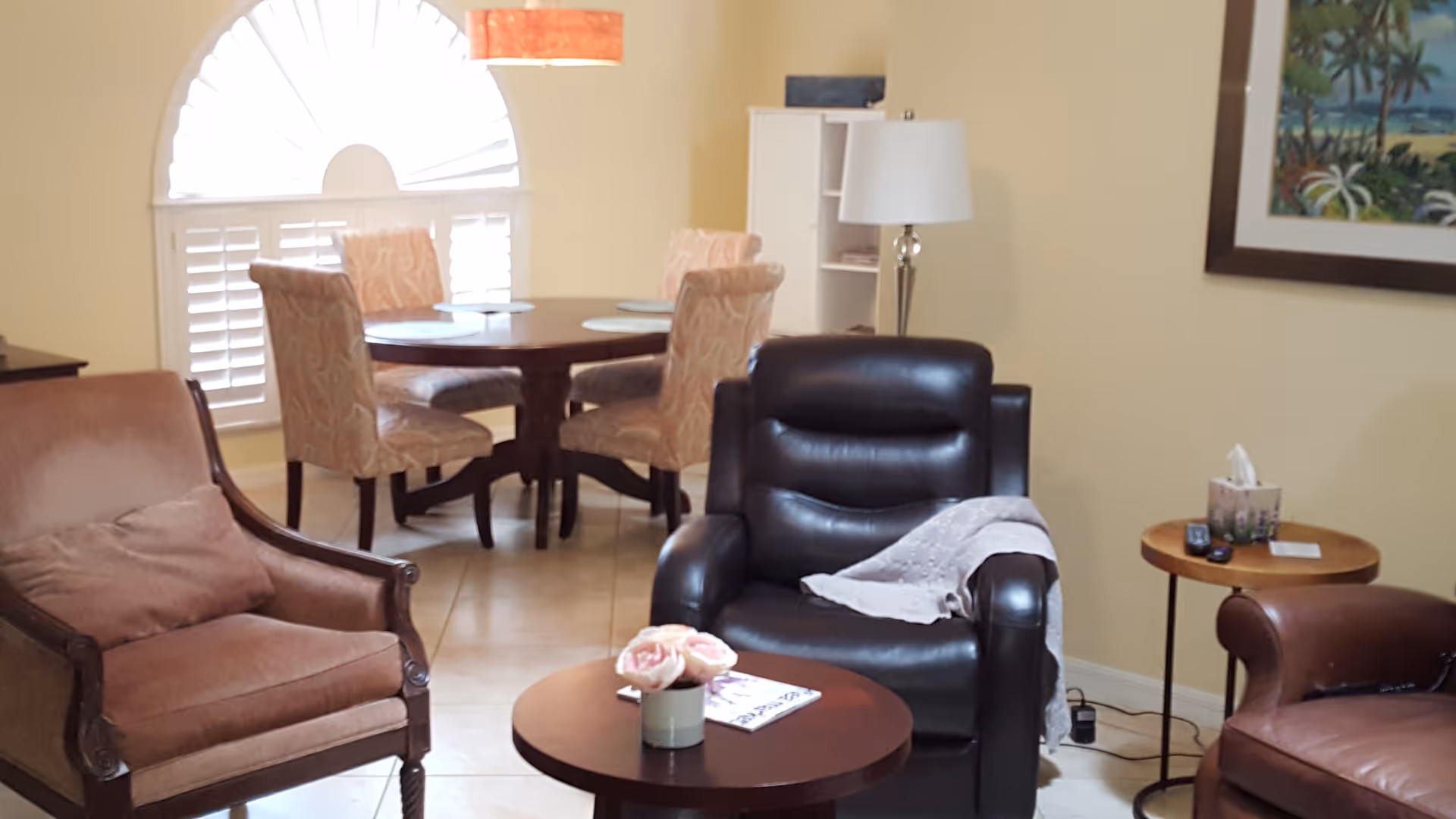 A cozy living room area with a brown leather armchair, a wooden armchair with a cushion, and a round wooden coffee table with a small flower arrangement and a magazine. In the background, there is a dining area with a round table and four upholstered chairs near a window with white shutters. A floor lamp and a small side table with tissues and remote controls are also visible.