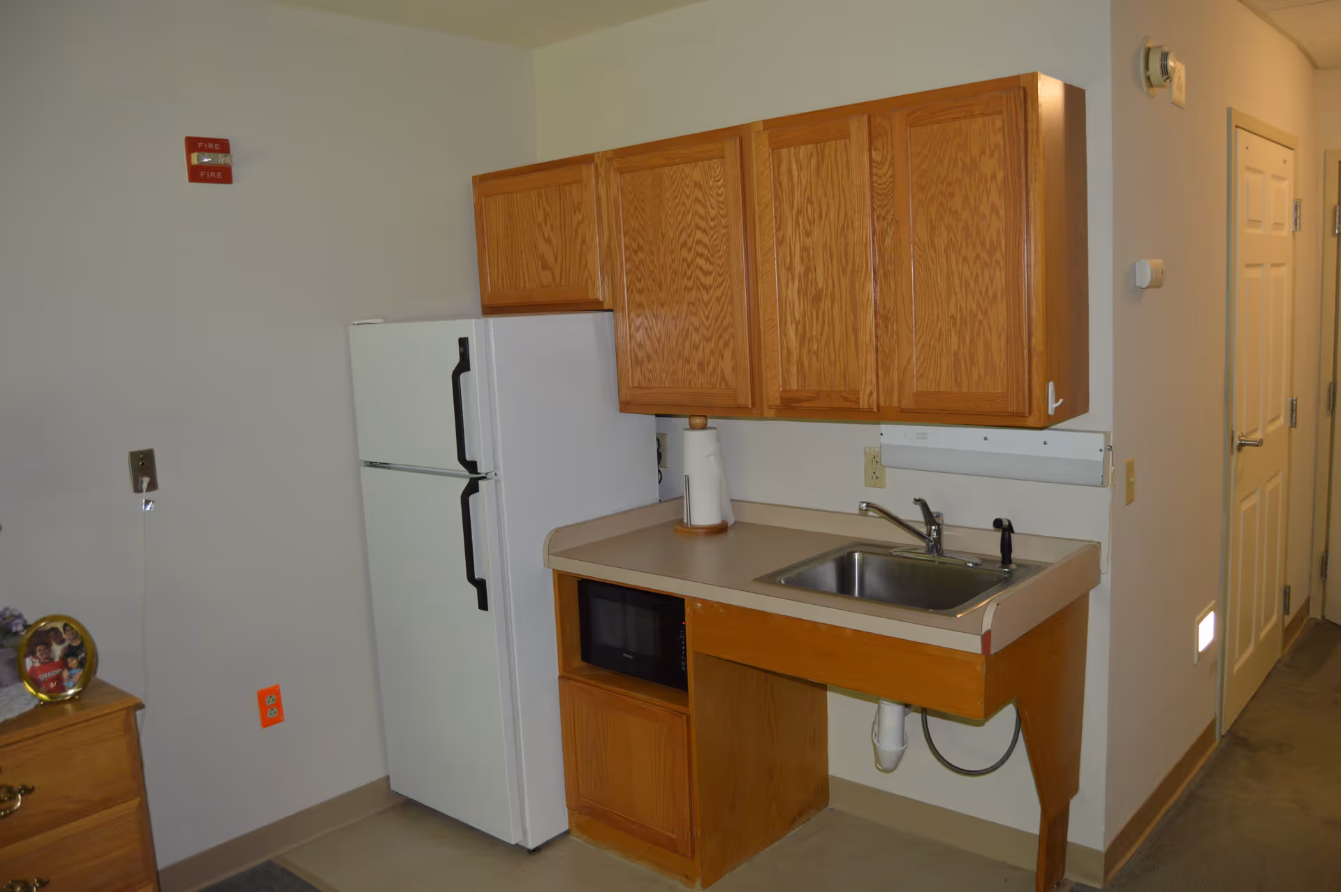 Small kitchen area with wooden cabinets, a white refrigerator, a microwave, a sink with a faucet, and a paper towel holder on the countertop. There is a fire alarm on the wall and a door visible in the background.