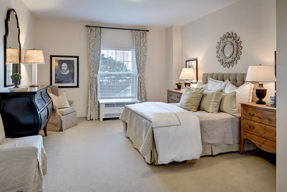 A well-lit bedroom with a large bed adorned with multiple pillows and a white comforter. On either side of the bed are wooden nightstands with lamps. A decorative round mirror hangs above the bed. To the left, there is a black dresser with a lamp and a small plant, a framed portrait on the wall, and a cushioned armchair with a pillow. A large window with patterned curtains lets in natural light, and the room has beige carpeting and light-colored walls.