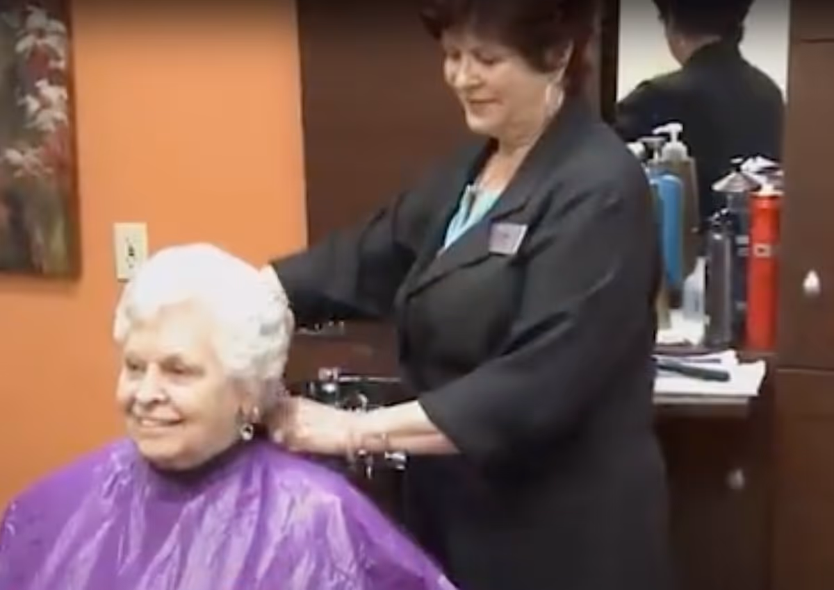 An elderly woman with white hair wearing a purple cape is sitting and smiling while a hairstylist stands behind her, styling her hair in a salon setting with orange walls and a mirror.