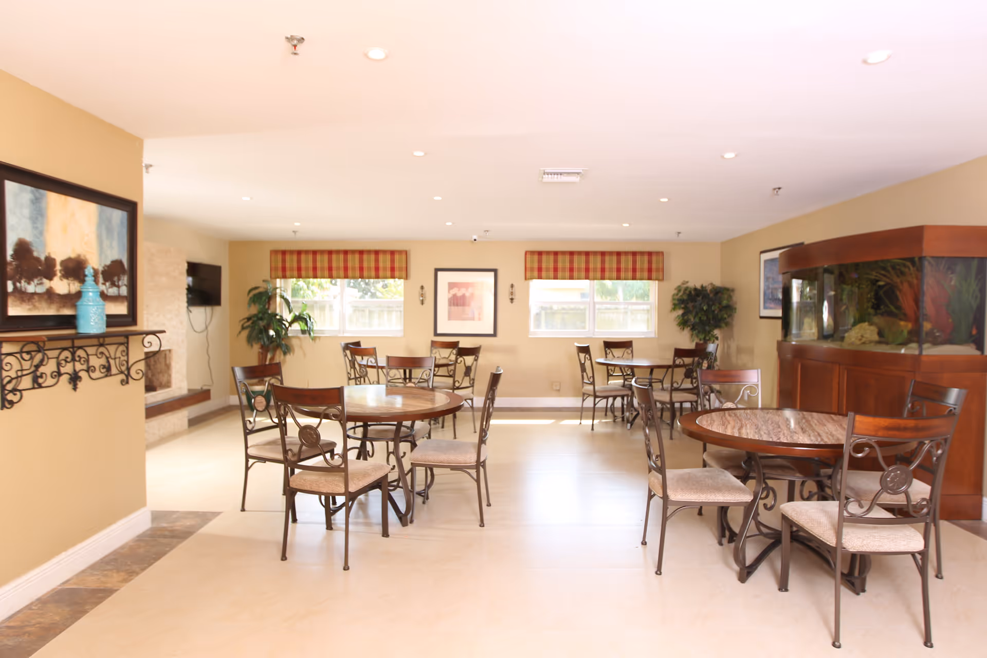 A bright and spacious dining room with several round tables and chairs arranged neatly. The room has beige walls, two windows with red and yellow plaid valances, framed artwork on the walls, and a large fish tank on the right side. There is also a decorative shelf with a blue vase and a wall-mounted TV near a fireplace on the left side.