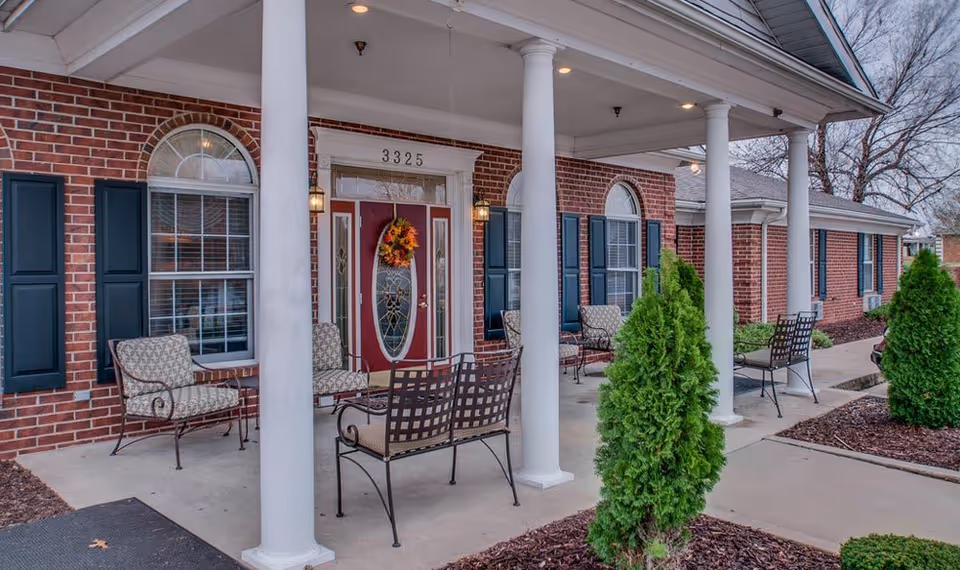Front porch of a brick building with white columns, several cushioned metal chairs and benches, a red door decorated with a fall wreath, and windows with dark blue shutters.