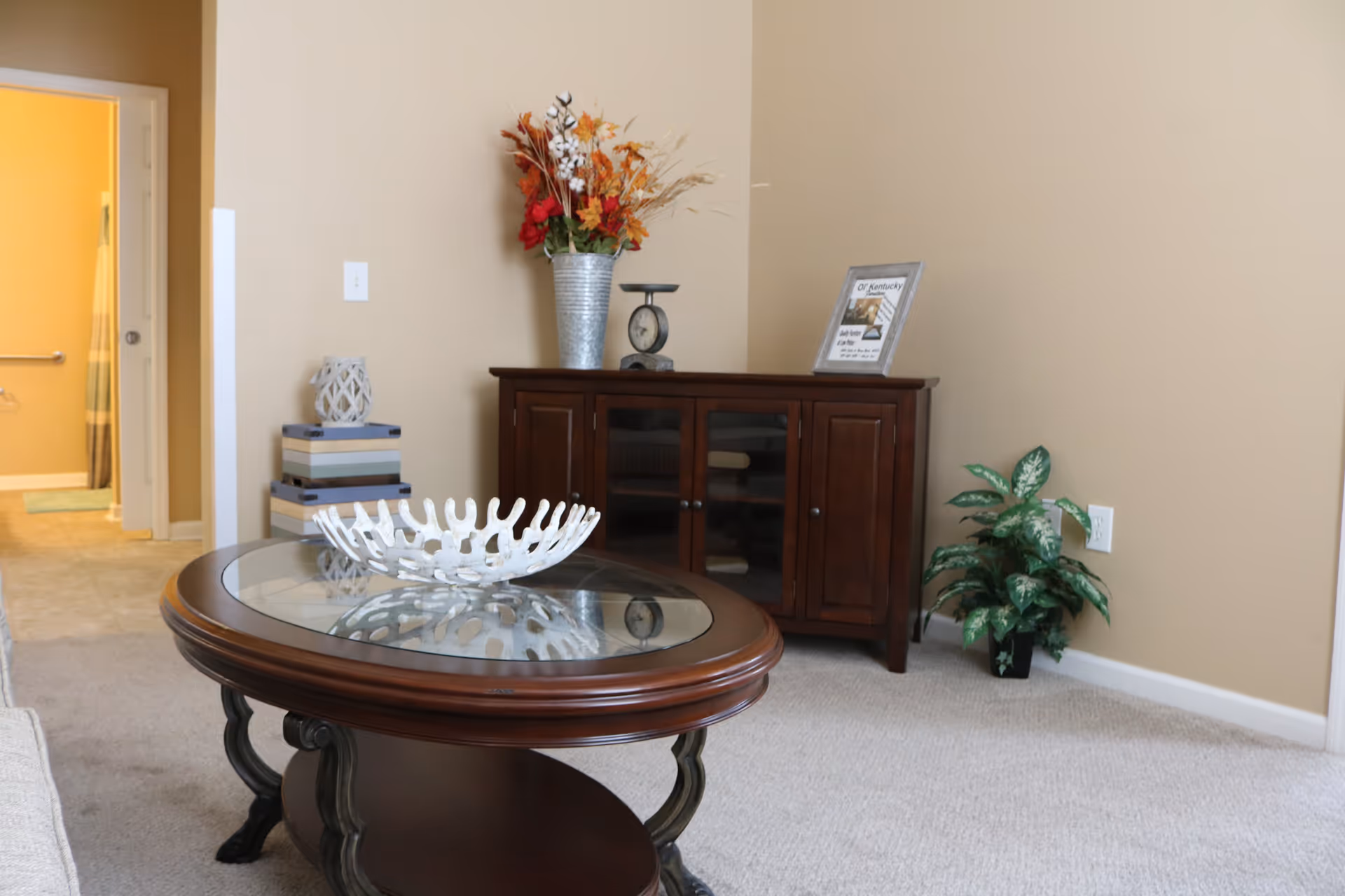 A cozy living room area with a round wooden coffee table featuring a decorative white bowl on top. Behind the table is a dark wooden cabinet with glass doors, decorated with a metal vase holding autumn-themed flowers, a vintage scale, and a framed picture. To the right of the cabinet is a green potted plant. The walls are beige, and there is a doorway leading to a bathroom with a shower curtain visible.