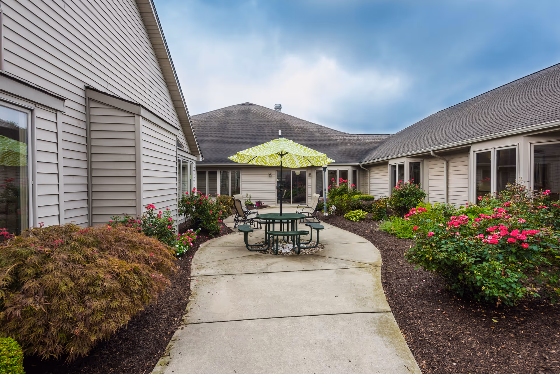 Courtyard with a concrete walkway leading to a patio table and green umbrella, surrounded by flowering shrubs and single-story beige buildings.