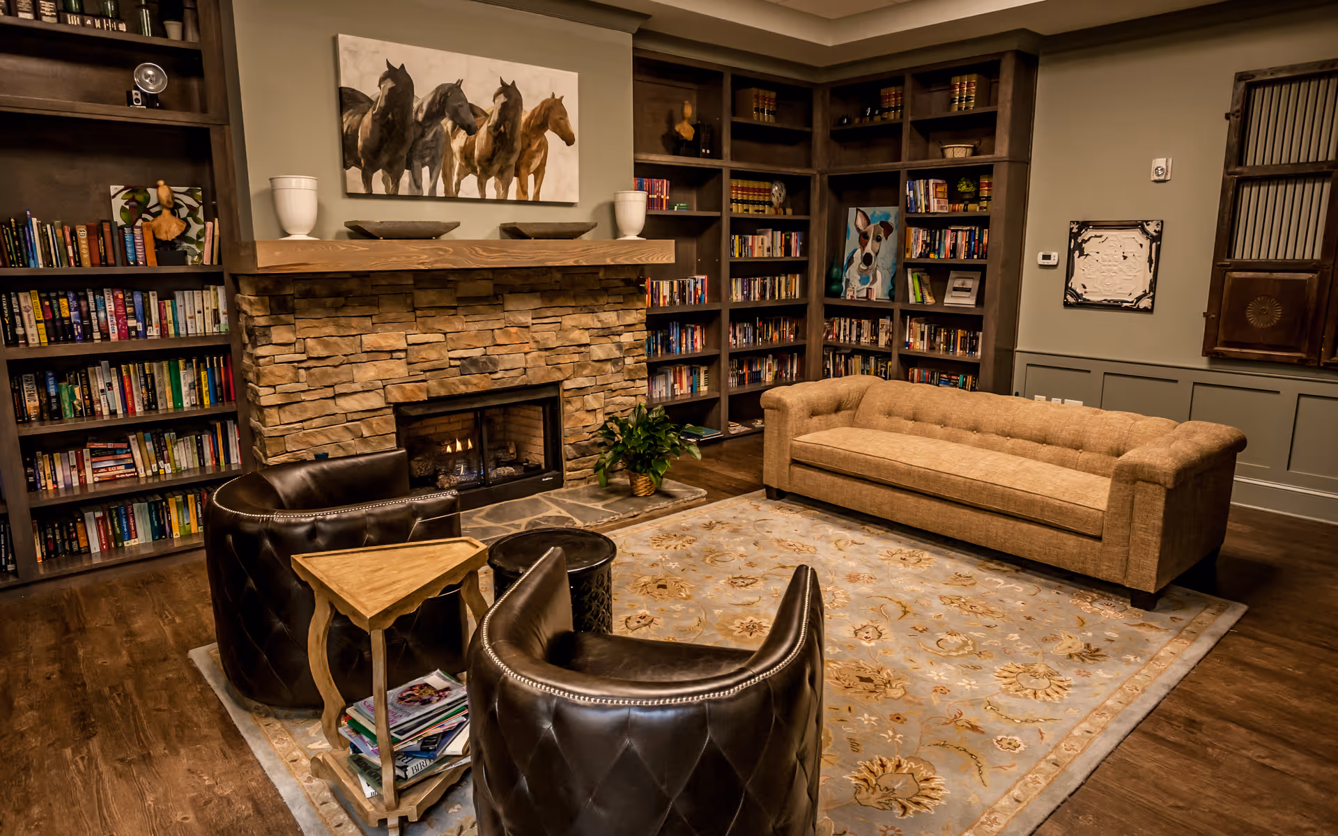 A cozy living room with a stone fireplace, a beige sofa, two dark leather armchairs, a wooden side table with magazines, and built-in bookshelves filled with books and decorative items. A painting of horses hangs above the fireplace.