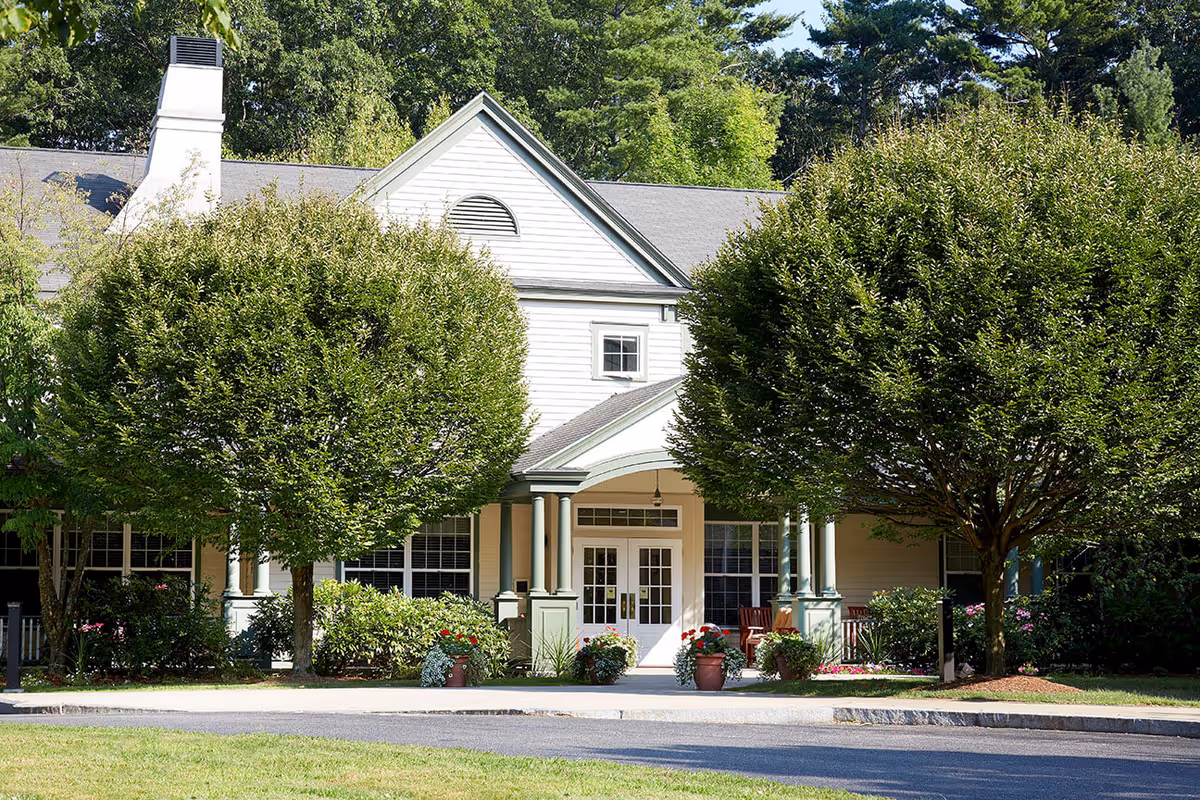 Front entrance of a two-story residential building with a covered porch, flanked by trimmed trees and potted flowers.