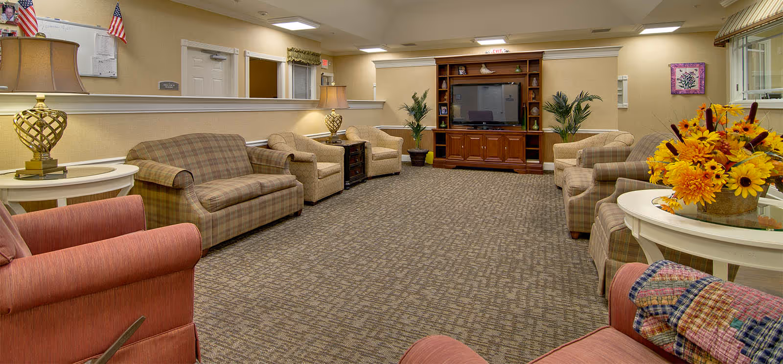 A spacious senior living facility common area with multiple upholstered armchairs and sofas arranged around the room. There are two side tables with decorative lamps, a large wooden entertainment center with a television, potted plants, and a round table with a bright floral arrangement. The walls are beige with framed artwork and an American flag is visible in the background.