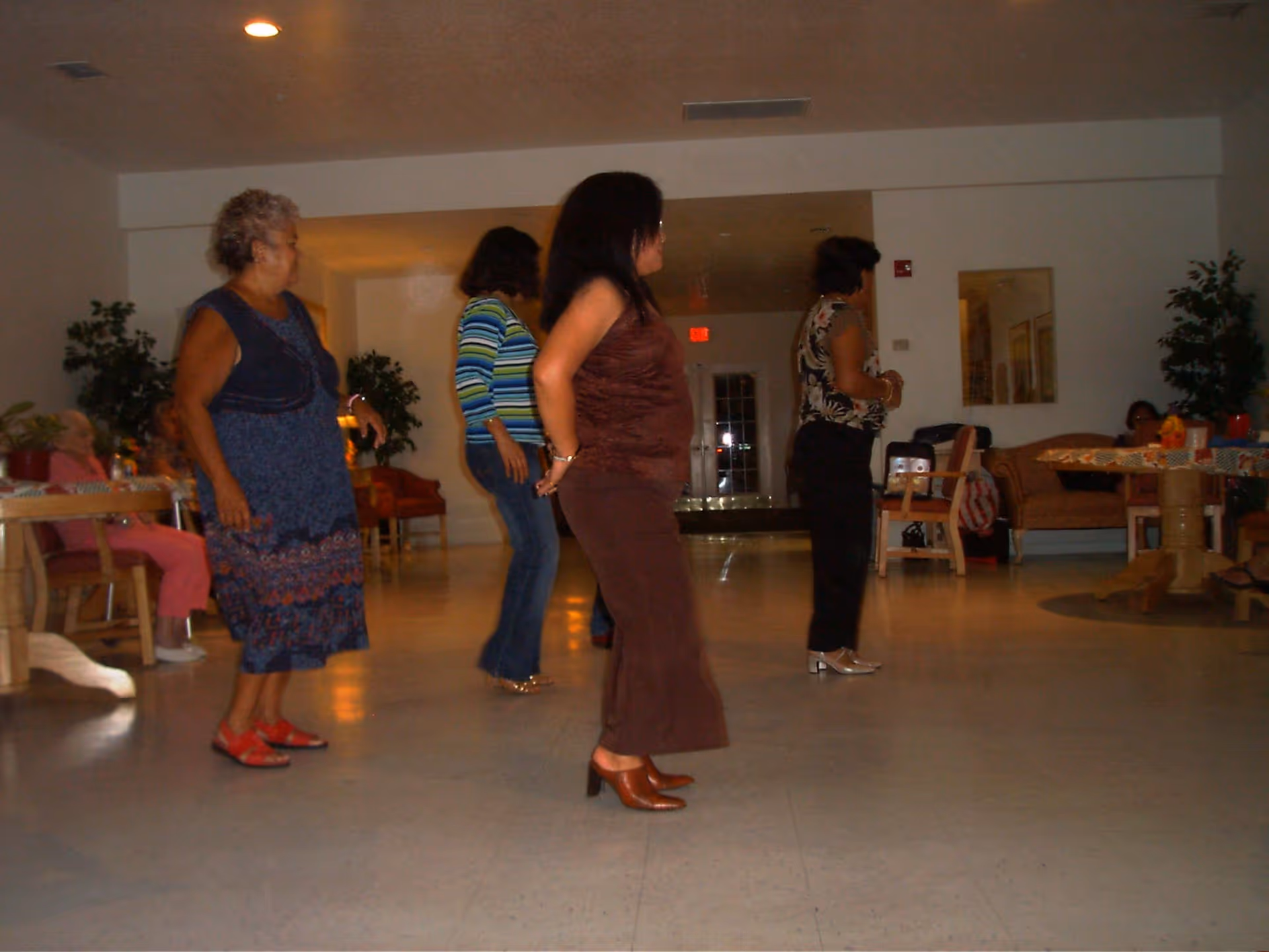 A group of four women standing and dancing in a spacious room with chairs, tables, and plants. The room has a polished floor and white walls with ceiling lights. Some people are seated in the background near tables.