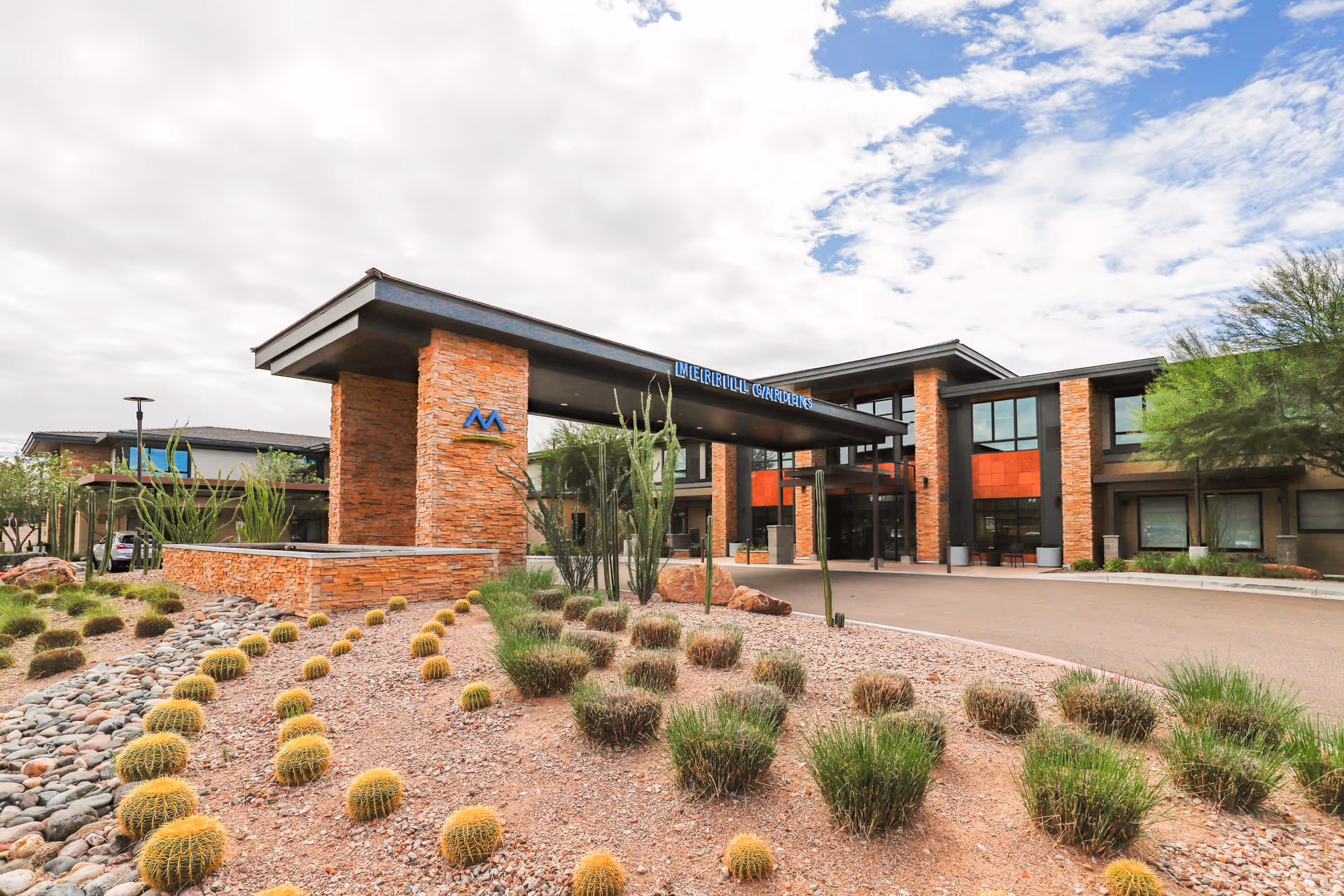 Exterior view of Merrill Gardens at Anthem senior living facility showing a modern building with stone pillars, large windows, and a covered entrance. The foreground features desert landscaping with cacti, rocks, and shrubs under a partly cloudy sky.