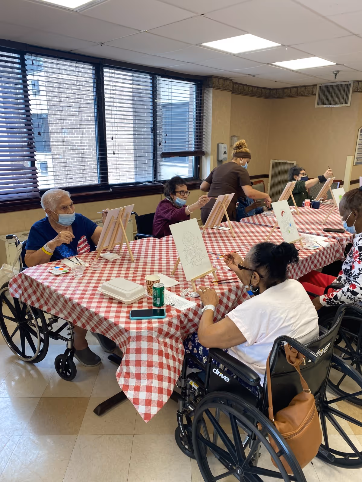 A group of elderly individuals, some in wheelchairs, seated around a long table covered with a red and white checkered tablecloth, participating in a painting activity. Each person has a canvas on an easel and is painting while wearing face masks. A caregiver is assisting one of the participants. The room has large windows with blinds and beige walls.