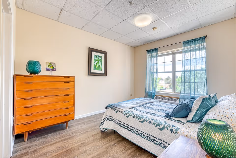 A bright bedroom with a large window covered by sheer blue curtains. The bed is made with a white and blue patterned quilt and several pillows. There is a wooden dresser with a green decorative vase and a small framed picture on top. A framed botanical print hangs on the wall above the dresser. The floor is wood, and the ceiling has white tiles with a round ceiling light.