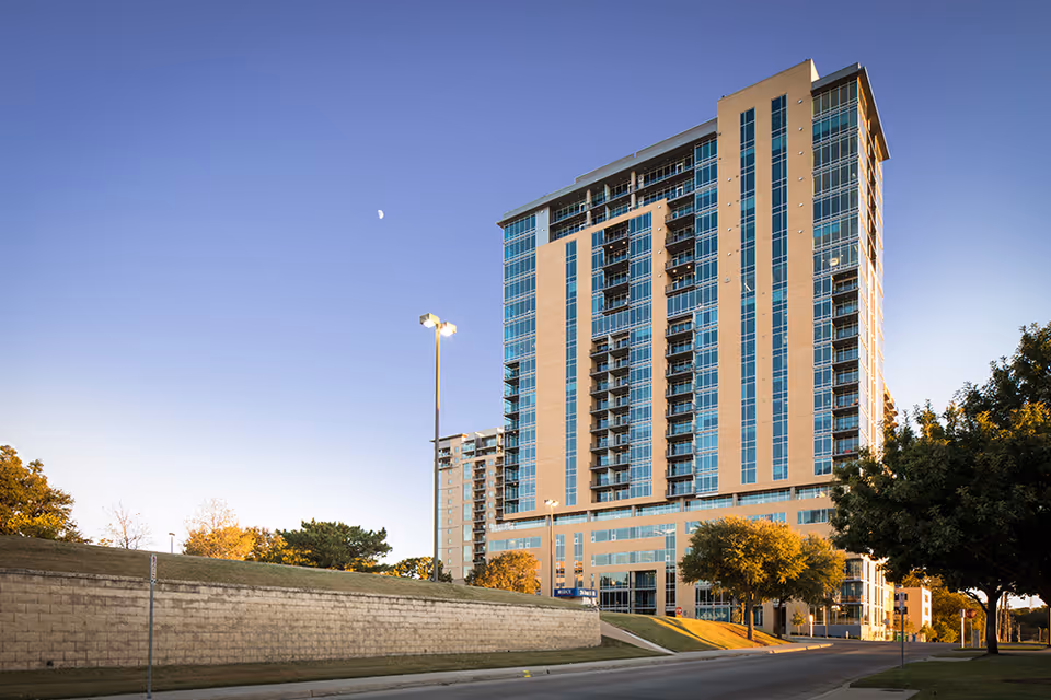 Tall modern building with many windows and balconies under a clear blue sky with a visible moon, surrounded by trees and a street in the foreground.