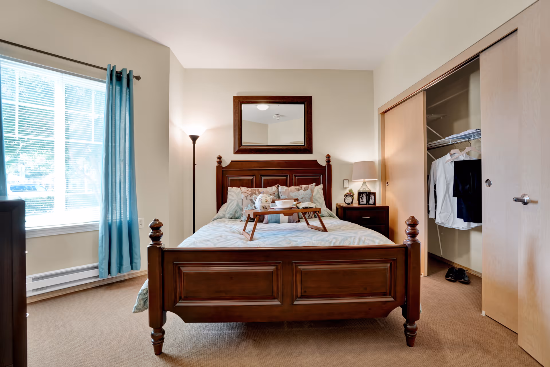 A bedroom with a wooden bed frame and a neatly made bed with patterned bedding. A wooden tray with a teapot and cup is placed on the bed. There is a large window with blue curtains on the left side, a floor lamp in the corner, and a nightstand with a lamp and framed photos on the right. A closet with sliding doors is open, showing hanging clothes and shoes inside. A large mirror hangs on the wall above the bed.