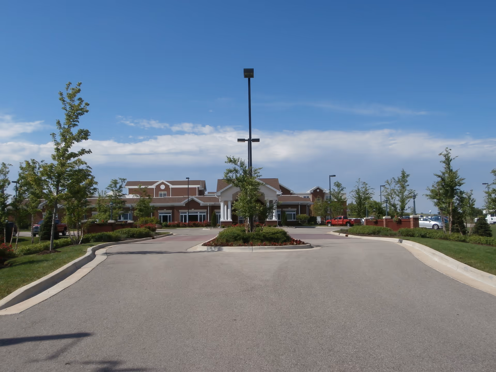 Front exterior view of Heritage Woods of Belvidere facility with a driveway leading to the entrance, landscaped greenery, trees, and a clear blue sky.